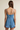 A woman shown from the back wearing a light blue denim romper with thin spaghetti straps and a ruffled hem, set against a plain beige studio background.