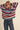 A woman wearing a chunky knit sweater with horizontal stripes in red, blue, green, pink, and cream, paired with dark blue jeans, posing with her hands behind her head against a solid beige background.