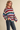 A woman wearing a fuzzy, oversized crewneck sweater with wide horizontal stripes in red, green, blue, cream, and pink, paired with dark blue jeans, set against a solid beige background.