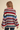 A woman shown from the back wearing a loose, oversized knit sweater with horizontal stripes in red, blue, green, cream, and pink, paired with dark blue jeans against a solid beige background.