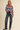 A woman modeling a fuzzy, oversized crewneck sweater with horizontal stripes in red, green, blue, pink, and cream, paired with dark blue jeans and dark flats, against a solid beige background.