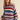 A woman wearing a chunky knit crewneck sweater with wide horizontal stripes in red, green, blue, pink, and beige, paired with dark jeans against a solid beige background.