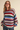 A woman wearing a chunky knit crewneck sweater with wide horizontal stripes in red, green, blue, pink, and beige, paired with dark jeans against a solid beige background.