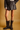 A model from the waist down wears a dark brown faux-leather mini skirt with front buttons and dark brown mid-calf boots with buckle straps, set against a solid warm tan studio background.