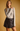 A woman modeling a light gray ribbed long-sleeve top and a dark brown faux-leather mini skirt against a solid warm tan background.