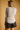 A woman shown from the back wearing a light beige ribbed knit long-sleeve top and dark brown faux-leather shorts, set against a solid warm tan background.