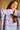 A woman wearing a chunky knit sweater with horizontal stripes in light blue, white, and red, posed against a solid warm tan background.