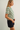 A woman in side profile wearing a green and white striped short-sleeve crop top and dark denim shorts, standing against a plain light gray studio background.