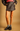 A model wearing a dark brown textured faux-leather mini skirt with side buttons, styled with a striped top, black leather jacket, gray socks, and black loafers against a solid tan background.