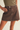A model wearing a dark brown denim mini skirt with a button closure and belt loops, paired with an olive green ribbed top, shown against a light beige background.