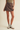 A model wearing a brown denim A-line mini skirt, styled with a ribbed olive green top, gray socks, and white sneakers with brown soles, set against a plain beige studio background.