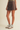 A model wearing a dark brown washed denim mini skirt, styled with a ribbed olive green top, gray socks, and white sneakers with black stripes, set against a plain beige studio background.