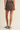 A model from the back wearing a brown washed denim mini skirt, paired with gray socks and white sneakers with navy stripes, against a solid light beige background.