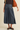A model is shown from the waist down wearing a dark blue denim midi skirt with a gathered waist, paired with a dark brown top, brown suede flats, and carrying a large brown suede shoulder bag against a warm off-white background.
