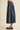 A person shown from the waist down wearing a dark blue denim midi skirt with gathered pleats and brown suede flats, standing against a plain beige studio background.