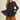 A woman modeling a black textured long-sleeve button-up blouse with a peplum waist, paired with wide-leg brown pants, against a solid beige background.