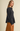 A woman wearing a dark gray plaid button-up shirt with a peplum waist and long sleeves, paired with a rust-colored skirt, standing against a solid beige background.