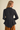 A woman shown from the back wearing a black textured long-sleeve peplum blazer and brown pants, set against a plain beige studio background.