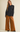 A woman modeling a black textured long-sleeve button-up shirt with a peplum waist, paired with wide-leg rust-brown pants and black shoes, against a solid light beige background.