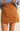 A close-up of a model wearing a tan faux-suede A-line mini skirt paired with a light blue knit sweater, shown from the waist down against a solid light beige background.