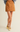 A model wearing a mustard yellow faux-leather mini skirt, styled with a gray knit sweater and yellow Mary Jane shoes against a warm beige studio background.