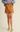 A model wearing a brown faux-leather A-line mini skirt, styled with a blue-gray knit sweater and pale yellow Mary Jane flats, against a solid beige studio background.