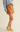 A model is shown from the waist down wearing a mustard brown suede mini skirt, a light blue knit sweater, and pale yellow Mary Jane shoes against a solid cream background.