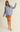 A woman modeling an oversized light blue-gray knit sweater with front patch pockets, paired with a brown mini skirt and pale yellow Mary Jane flats, against a plain beige studio background.