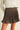 A brown pleated mini skirt with a matching belt and gold buckle, styled with a white ribbed sweater on a model.