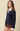 A woman modeling a navy blue knit sweater layered over a white collared shirt with brown shorts, set against a solid beige studio background.