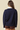 A woman shown from the back wearing a navy blue knit sweater over a white collared shirt, paired with brown shorts, against a solid beige background.