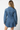 A woman viewed from behind wearing a medium-wash blue denim shirt jacket with long sleeves and a relaxed fit, set against a plain white studio background.