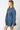 A woman wearing a blue denim long-sleeve shirt dress, shown from a side profile against a plain white studio background.