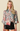 A woman modeling a long-sleeve satin blouse with a large floral print in pink, red, green, and beige tones on a light gray base, paired with black shorts, against a solid beige background.