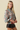 A blonde model wearing a long-sleeve floral blouse with a high mock neck and black faux leather shorts, posed against a solid beige studio background.
