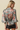 A woman shown from the back wearing a sheer gray satin blouse with a large pink and red floral print, paired with black shorts, against a solid beige background.