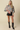 A blonde model stands against a plain beige studio background wearing a floral satin blouse with puff sleeves, black faux-leather shorts, and black pointed-toe heeled boots.