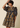 A woman wearing a navy blue tiered dress with a mustard yellow floral print, featuring puff sleeves and a ruffled neckline, posed against a solid beige background.