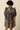 A model shown from the back wearing a navy blue tiered dress with a mustard yellow floral print, puff sleeves, and a ruffled collar, set against a solid beige background.