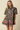A woman modeling a short-sleeve button-up shirt dress with a dark green base and a pink, red, and beige floral pattern, set against a solid beige background.