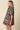 A woman wearing a dark green short-sleeve button-up dress with a pink and red floral print and a patterned hem, photographed against a plain beige studio background.