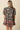 A woman viewed from the back wearing a dark green floral puff-sleeve mini dress with a patterned hem, set against a solid beige background.