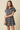 A young woman modeling a short-sleeve plaid shirt dress with a button-down front and flared skirt, set against a solid beige studio background.