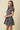 A young woman modeling a short-sleeve plaid mini dress with a button-up front and flared skirt, photographed against a plain beige studio background.