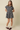 A young woman modeling a short-sleeve plaid mini dress with a button-up front and flared skirt, paired with cream socks and burgundy loafers, against a solid beige studio background.