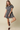 A woman modeling a short-sleeve plaid mini dress with a button-up front and flared skirt, paired with cream socks and dark brown loafers, against a solid beige studio background.