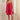 A woman modeling a bright pink pleated slip dress with thin spaghetti straps, standing in a classic indoor room with white walls, a wooden floor, and a cream curtain.