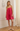 A woman modeling a bright pink pleated slip dress with thin spaghetti straps, standing in a classic indoor room with white walls, a wooden floor, and a cream curtain.
