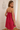 A woman modeling a bright pink and red vertically striped slip dress with thin spaghetti straps, shown from a three-quarters back view in an elegant indoor setting with a white wall and wooden floor.