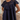 A woman wearing a navy blue short-sleeve tunic top with a ruffled yoke and embroidered detailing, shown in a softly lit indoor setting.
